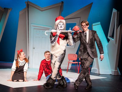 The Cat in the Hat is holding an umbrella like a flute. There is a man with a puppet holding the brolly in its mouth. A girl and a boy sit on the floor by their feet