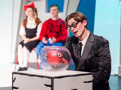 a man in a black suit and glasses looks at a fish bowl with a red fish puppet inside