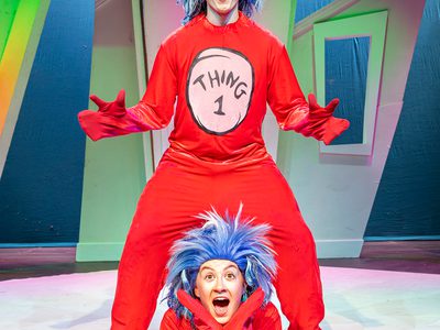 Thing 1 in a red suit with blue wig stands over Thing 2 dressed the same who lays in between his legs