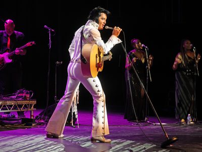 Ben Portsmouth as Elvis wears a tight white bejewelled jumpsuit leans forward as he sings into a microphone