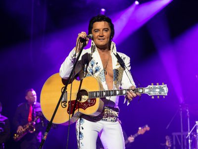 Ben Portsmouth as Elvis wears a tight white bejewelled jumpsuit and he smiles as he holds a guitar and microphone