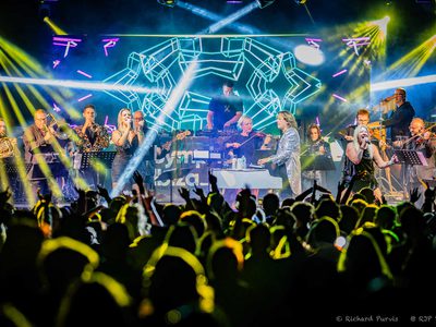 A stage full of people with beams of yellow and blue lights covering the stage and crowd. The stage has people singing, DJing, and playing various instruments