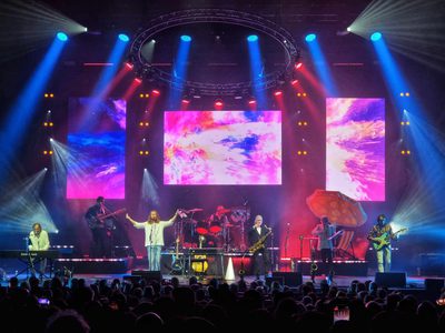 A vibrant rock concert scene with colourful stage lights. The band performs energetically under a circular light rig and abstract pink visuals on screens
