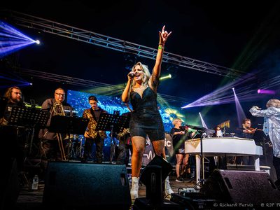 A woman in front of the orchestra in a black dress singing into a microphone and holding her hand in the air doing the rock horn sign with her fingers