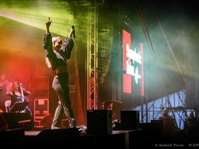 A woman walking out towards the front of the stage with red and green lights behind her holding her arms in the air with a microphone in one hand