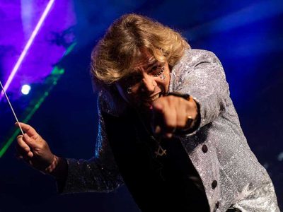The conductor of the orchestra ina silver jacket with gems on his face smiling at and pointing at the camera while holding his baton in the other hand