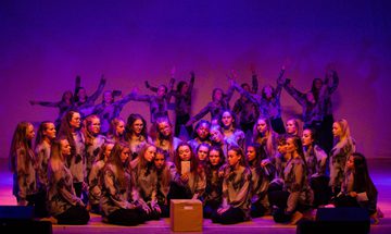 a group of girl dancers knelt on the floor. The one at the front in the middle is holding up a letter with a wooden box in front