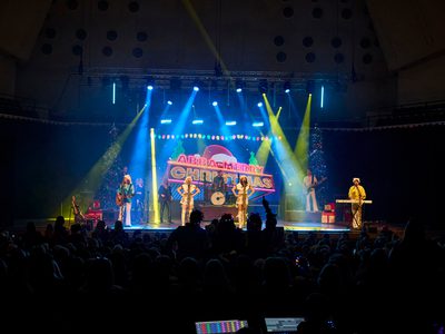 A band performs on stage in front of a live audience. The two lead singers at the front are wearing white dresses and Santa hats. The band also includes a keyboard player, two guitarists, and background singers. Bright blue and green lights illuminate the stage.