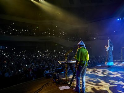 A photo taken from behind a keyboard player on stage. The keyboard player is wearing a green shirt, and the audience is facing the stage.