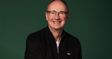 Jonathan Agnew, a smiling man with glasses wearing a black zip-up jacket, against a dark green background