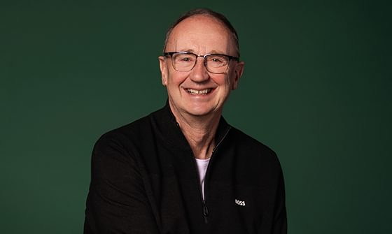 Jonathan Agnew, a smiling man with glasses wearing a black zip-up jacket, against a dark green background