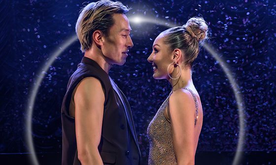 Carlos wears his hair slicked back and a black sleeveless vest. Amy faces him wearing a sparkly dress and her hair in a high bun.