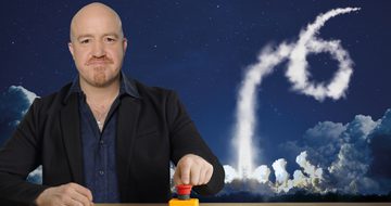 Andy Parsons a bald, middle-aged man sits at a wooden desk in a black suit. On the desk is a big red button which he has his finger on. Behind him are vapor trails in the sky