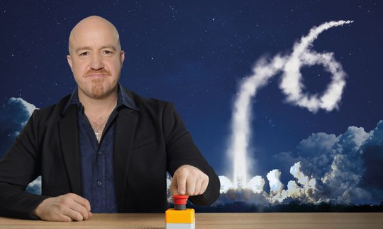 Andy Parsons a bald, middle-aged man sits at a wooden desk in a black suit. On the desk is a big red button which he has his finger on. Behind him are vapor trails in the sky