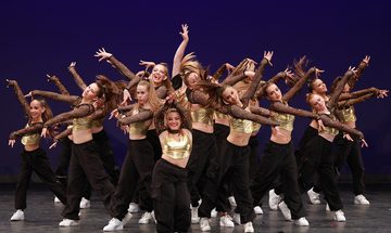 A group of girls in black trousers, white trainers, and gold tops with long net sleeves all in a large group posing with their arms stretched either up or out to the side