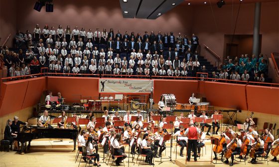 A large group of children in white shirts and various coloured ties at The Anvil performing on stage with instruments or singing in the choir stalls