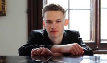Kasparas Mikuzis, a young man in a black suit sits with his back to a large window with his hands resting on the top of a piano