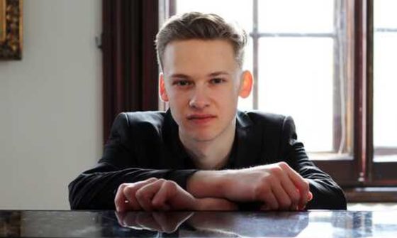 Kasparas Mikuzis, a young man in a black suit sits with his back to a large window with his hands resting on the top of a piano