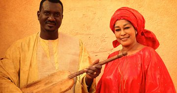 Bassekou a black man in a cream shirt, holding a ngoni, stood with Amy a black woman in a red dress with a matching head wrap