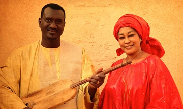 Bassekou a black man in a cream shirt, holding a ngoni, stood with Amy a black woman in a red dress with a matching head wrap