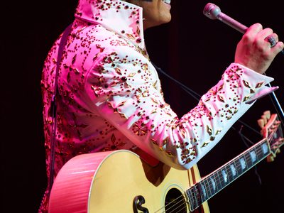 Ben Portsmouth as Elvis wears a tight white bejewelled jumpsuit and he smiles as he holds a guitar and microphone