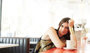 A woman with long brown hair, wearing a black and gold tank top and gold bangle, leans her elbow on the red countertop of a cafe, before her sits a cup of coffee
