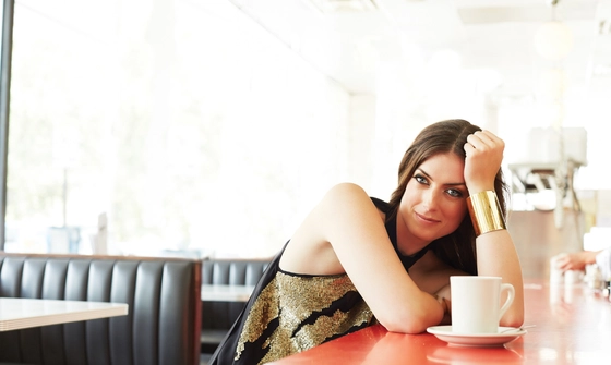A woman with long brown hair, wearing a black and gold tank top and gold bangle, leans her elbow on the red countertop of a cafe, before her sits a cup of coffee