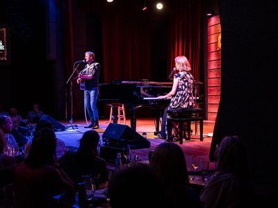 A side view of the stage from amongst the audience. On stage is a man playing guitar and singing and to his left is a woman on piano