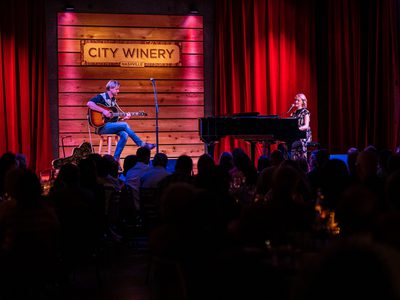 A man sits on a stool and plays an acoustic guitar while to his left a woman performs on the piano, behind them is a wooden wall and a sign reading 'City Winery'