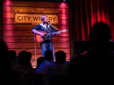 A man plays an acoustic guitar while singing into a microphone on a stand, behind him is a wooden wall and a sign reading 'City Winery'