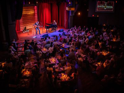 A large audience all watch as a man and a woman play guitar and piano on stage