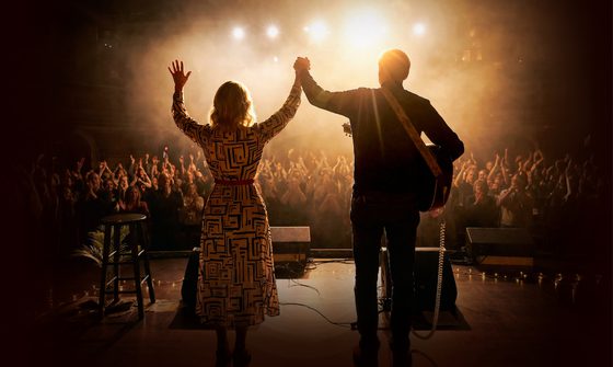 Carole King and James Taylor stand with their back to the camera but facing the audience as they cheer and clap at the end of a musical performance