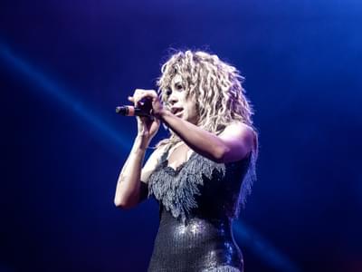 A Tina Turner tribute with curly hair holding a microphone. She is wearing a sparkly dress against a blue-lit background
