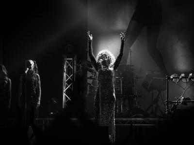 Black and white image of a singer on stage, arms raised, wearing a sparkling dress. Dimly lit atmosphere with band equipment in the background