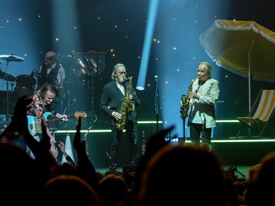 A lively concert scene with four musicians on stage, two playing saxophones and one on guitar, and a drummer is in the back