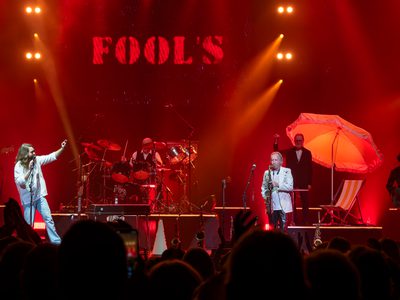A band performing under vibrant red and orange stage lights. A singer points passionately, surrounded by musicians playing instruments