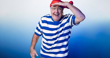 Danny Baker, a man wearing a red bandana, a blue and white striped shirt, and glasses, smiling and playfully posing with a hand raised above his eyes