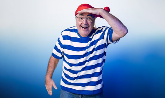 Danny Baker, a man wearing a red bandana, a blue and white striped shirt, and glasses, smiling and playfully posing with a hand raised above his eyes