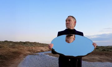 David Gray wears all black as he stands on a gravel path in the countryside, he holds a round mirror below his face, reflecting his face and the blue sky above