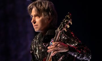 Eric looks focussed as he looks ahead. He's wearing a silvery shirt and his guitar has a colourful strap which rests on his shoulder.