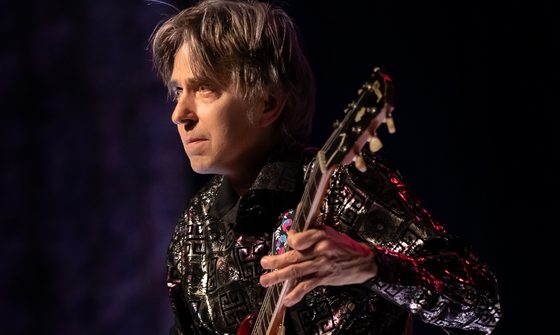 Eric looks focussed as he looks ahead. He's wearing a silvery shirt and his guitar has a colourful strap which rests on his shoulder.