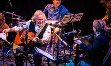 An old man sat on stage with long white hair and a short white beard holding a guitar surrounded by other band members all with various instruments, microphones, and music stands