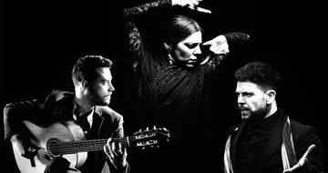 A black and white photo with three people, one man in a suit playing guitar, a woman flamenco dancing in a lacey dress, and another man wearing a scarf looking very angry