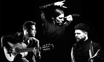 A black and white photo with three people, one man in a suit playing guitar, a woman flamenco dancing in a lacey dress, and another man wearing a scarf looking very angry