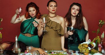 three women stood behind a table holding wine glasses. A fourth woman is passed out on the table