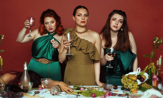 three women stood behind a table holding wine glasses. A fourth woman is passed out on the table