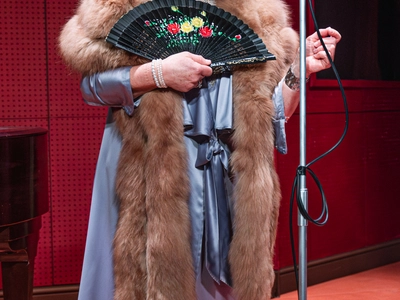 Wendi Peters, an older white woman with short brown hair, in a long sleeved, light blue, silk dress, with a large fur around her neck reaching her knees, and a small woven hat, holding a black, floral, hand fan. She is in a recording studio looking up towards a microphone