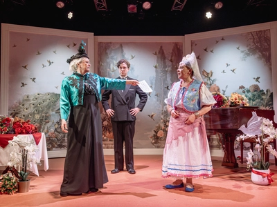 Wendi Peters, another older woman, and a young man on stage together. They're stood in a bright room with pictures of bird as walls, there is also a piano, a table, and dozens of flowers. The other woman, who is in a black dress, and hat with a turquoise jacket is hanging a letter to a confused looking Wendi Peters who is in an outfit reminiscant of traditional Scandinavian dress with a white skirt and blouse that have blue and pink detailing, a light blue vest with colourful patterns and a lace headdress.