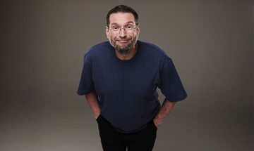 Gary Delaney in a blue t-shirt and black jeans with his hands in his pockets leaning forward and awkwardly smiling