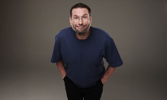 Gary Delaney in a blue t-shirt and black jeans with his hands in his pockets leaning forward and awkwardly smiling